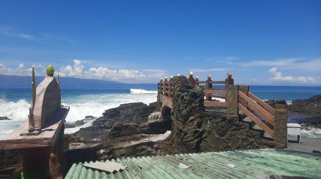 Sebuah struktur beton lengkung berdiri mencolok di antara karang-karang alami Pantai Karang Hawu, Kecamatan Cisolok, Kabupaten Sukabumi. Di bagian atasnya terukir tulisan "Alor Mandung" berwarna emas yang menarik perhatian para pengunjung.